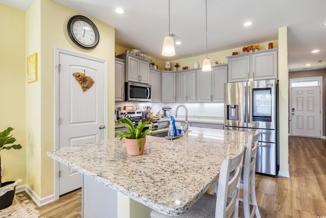 kitchen featuring gray cabinets, appliances with stainless steel finishes, hanging light fixtures, light stone countertops, and an island with sink