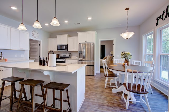 kitchen with an island with sink, wood-type flooring, decorative light fixtures, white cabinets, and appliances with stainless steel finishes