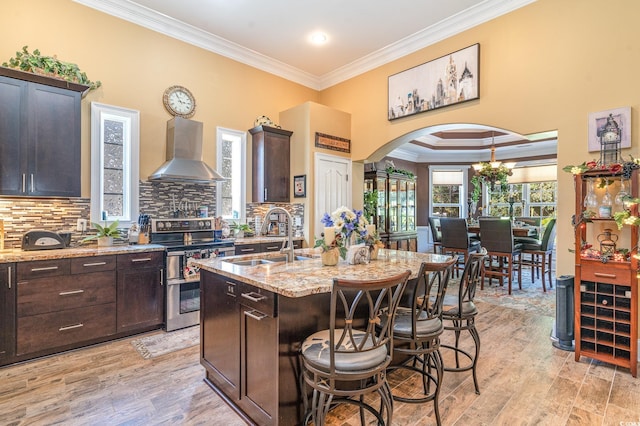 kitchen featuring wall chimney range hood, an island with sink, light stone countertops, light hardwood / wood-style floors, and electric stove