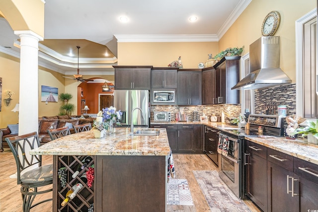 kitchen with an island with sink, decorative columns, a kitchen breakfast bar, stainless steel appliances, and wall chimney exhaust hood