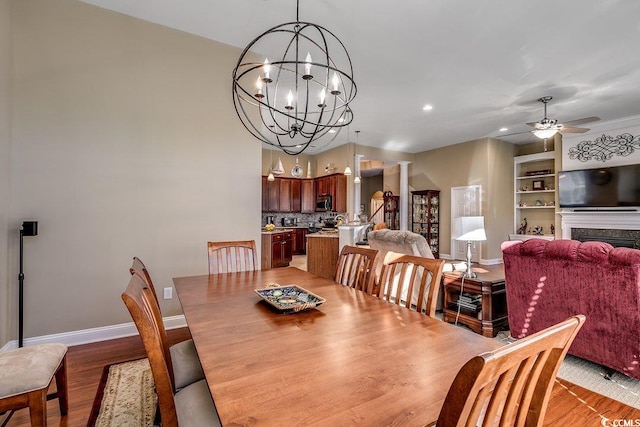 dining area featuring decorative columns, a fireplace, baseboards, ceiling fan with notable chandelier, and light wood finished floors