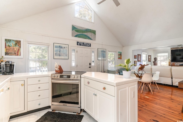 kitchen with tile countertops, high vaulted ceiling, stainless steel electric stove, white cabinets, and light wood-type flooring