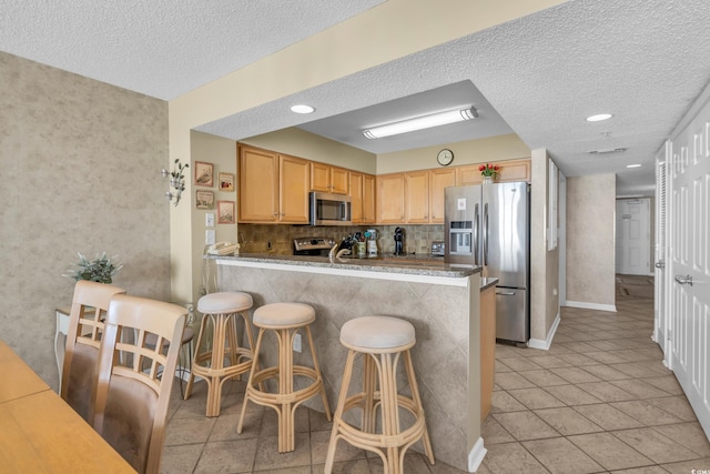 kitchen with stainless steel appliances, a textured ceiling, light brown cabinetry, a kitchen breakfast bar, and kitchen peninsula