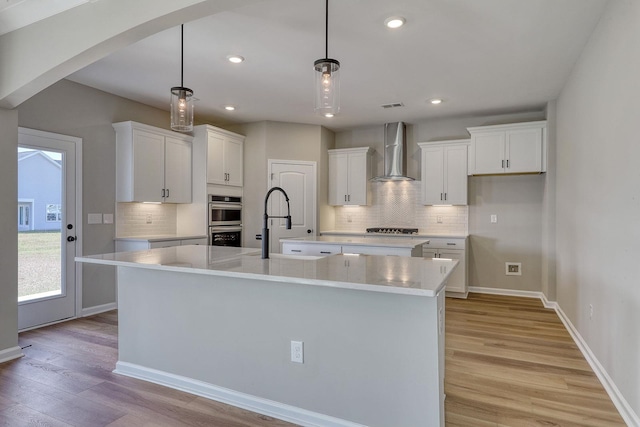 kitchen with a kitchen island with sink, hanging light fixtures, white cabinets, and wall chimney range hood