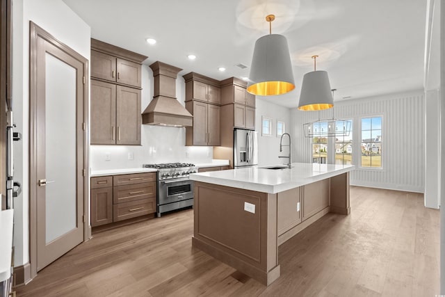 kitchen featuring appliances with stainless steel finishes, custom range hood, sink, decorative light fixtures, and a center island with sink