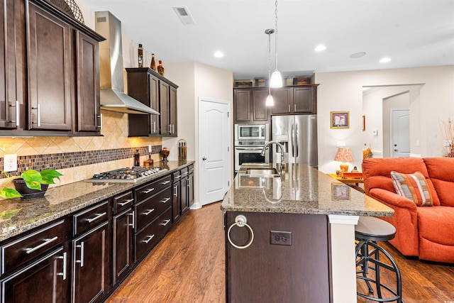 kitchen featuring stainless steel appliances, a center island with sink, a breakfast bar area, dark wood-type flooring, and wall chimney range hood