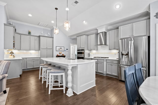 kitchen with appliances with stainless steel finishes, wall chimney exhaust hood, hanging light fixtures, a kitchen island with sink, and dark wood-type flooring