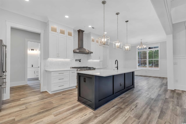 kitchen with white cabinetry, light countertops, wall chimney exhaust hood, an island with sink, and glass insert cabinets