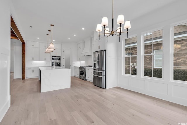 kitchen with appliances with stainless steel finishes, custom exhaust hood, a kitchen island with sink, decorative light fixtures, and white cabinetry