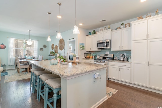 kitchen with hanging light fixtures, appliances with stainless steel finishes, dark hardwood / wood-style floors, and a kitchen island with sink