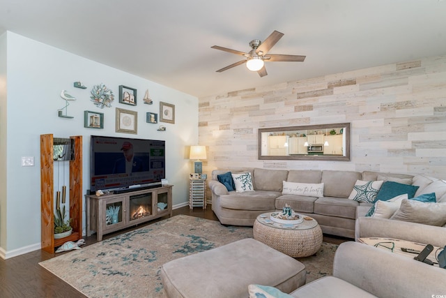 living area featuring a ceiling fan, a glass covered fireplace, wood finished floors, baseboards, and an accent wall