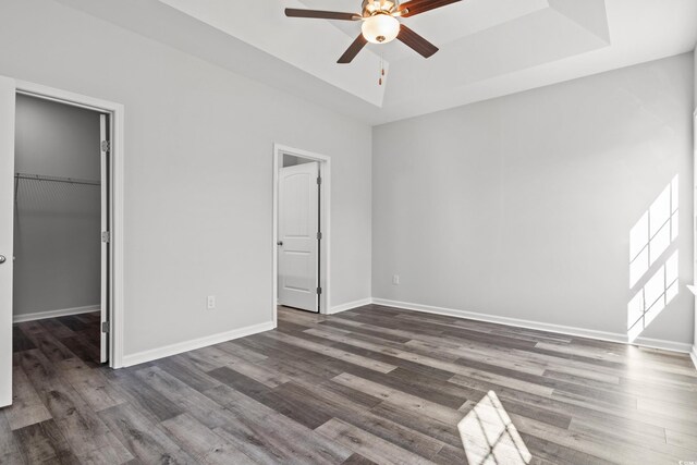 unfurnished bedroom with a walk in closet, a tray ceiling, dark hardwood / wood-style flooring, a closet, and ceiling fan