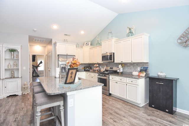 kitchen with a kitchen bar, vaulted ceiling, an island with sink, stainless steel appliances, and decorative backsplash