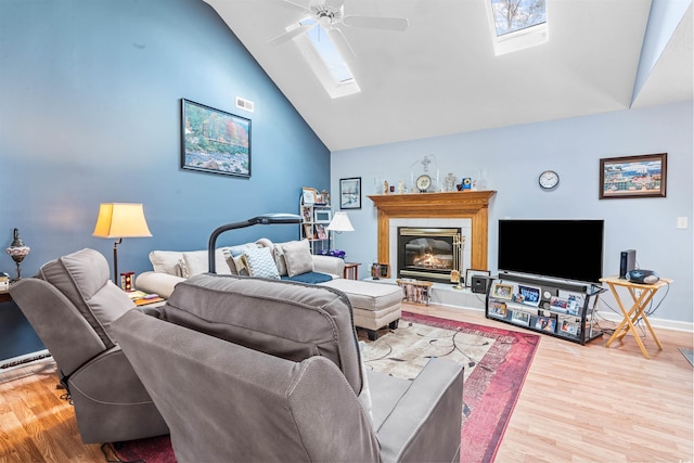living area featuring a skylight, wood finished floors, a ceiling fan, high vaulted ceiling, and a glass covered fireplace