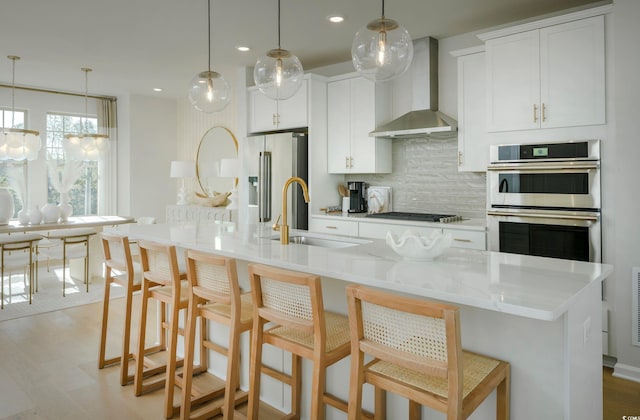 kitchen with stainless steel appliances, a kitchen island with sink, sink, wall chimney range hood, and hanging light fixtures