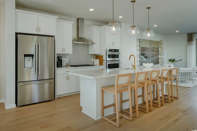 kitchen with white cabinets, wall chimney range hood, hanging light fixtures, an island with sink, and appliances with stainless steel finishes