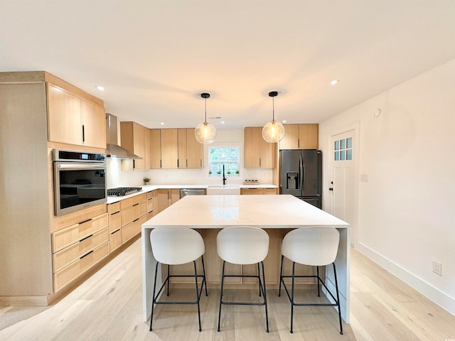 kitchen with stainless steel appliances, wall chimney range hood, a kitchen island, light brown cabinetry, and decorative light fixtures