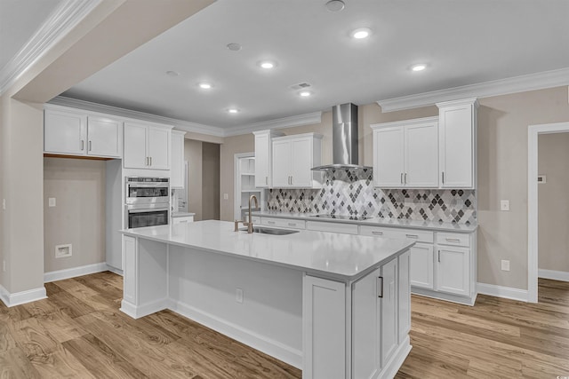 kitchen featuring white cabinetry, sink, wall chimney exhaust hood, stainless steel double oven, and an island with sink