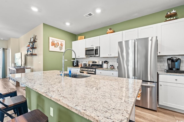 kitchen featuring light stone countertops, light wood-type flooring, stainless steel appliances, a center island with sink, and white cabinets