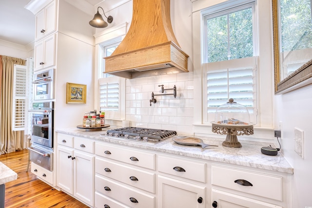 kitchen featuring appliances with stainless steel finishes, premium range hood, light stone countertops, light hardwood / wood-style floors, and white cabinets