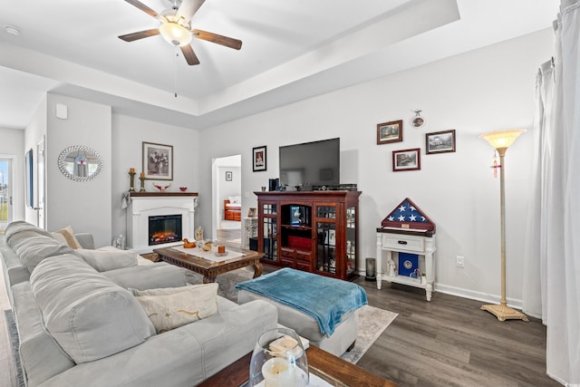 living area featuring baseboards, a raised ceiling, a ceiling fan, dark wood-type flooring, and a glass covered fireplace