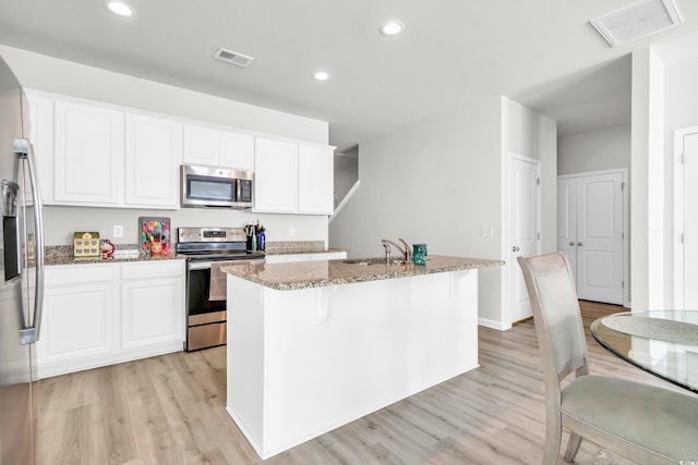 kitchen with a kitchen island with sink, light stone countertops, appliances with stainless steel finishes, light hardwood / wood-style floors, and white cabinetry