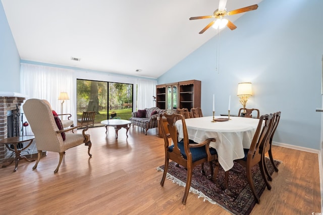 dining area featuring a brick fireplace, light wood-type flooring, high vaulted ceiling, and ceiling fan