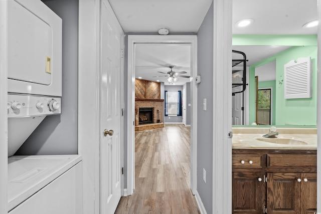 laundry room with sink, light hardwood / wood-style flooring, a brick fireplace, ceiling fan, and stacked washing maching and dryer