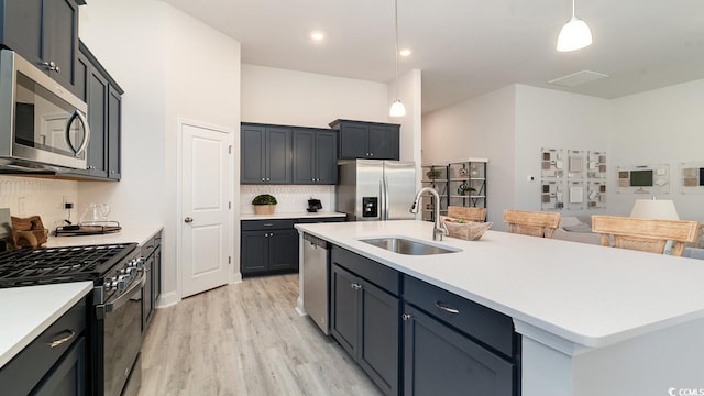 kitchen with light wood-type flooring, stainless steel appliances, sink, a center island with sink, and hanging light fixtures