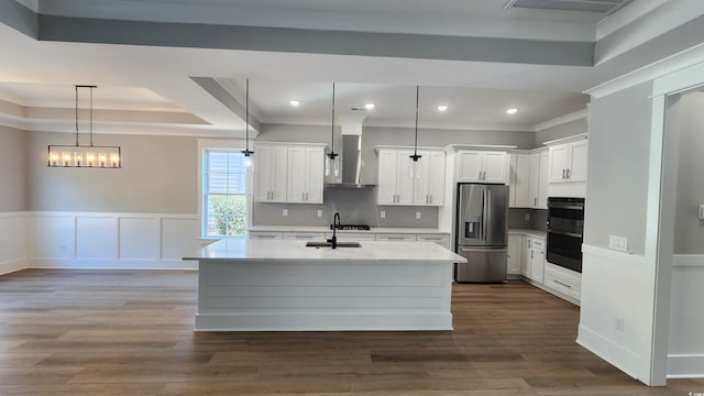 kitchen featuring a center island with sink, stainless steel refrigerator with ice dispenser, a raised ceiling, hanging light fixtures, and wall chimney range hood