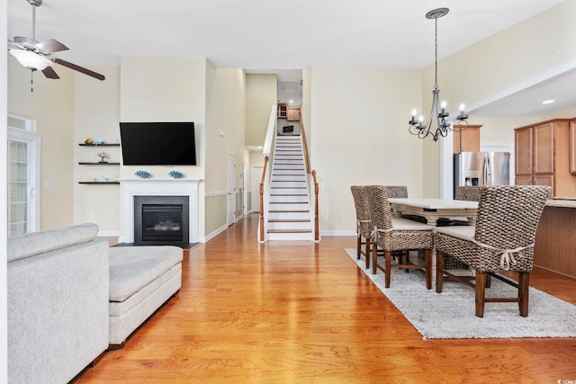 dining room with stairs, light wood-style floors, a fireplace with flush hearth, baseboards, and ceiling fan with notable chandelier