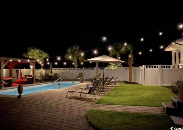 pool at night featuring a patio area, a fenced backyard, a yard, and a community pool