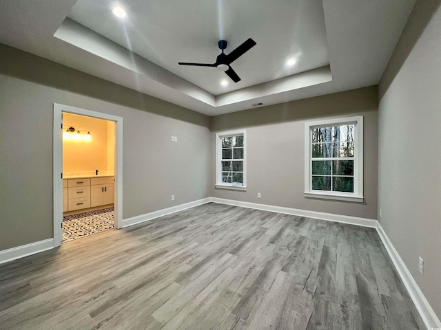 unfurnished bedroom featuring ensuite bath, ceiling fan, hardwood / wood-style floors, a tray ceiling, and multiple windows