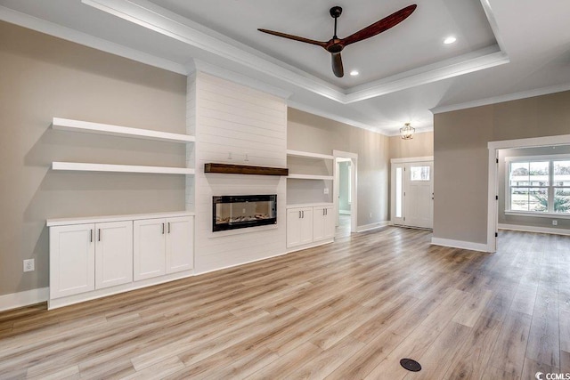 unfurnished living room with a tray ceiling, a multi sided fireplace, ornamental molding, ceiling fan with notable chandelier, and built in shelves