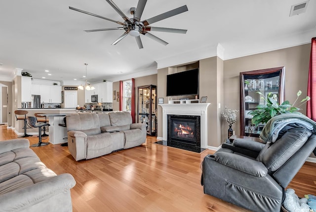 living room featuring ceiling fan with notable chandelier, light hardwood / wood-style floors, crown molding, and a high end fireplace