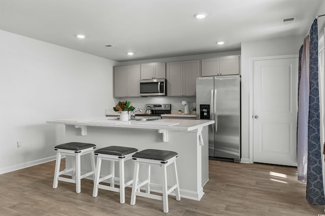 kitchen with gray cabinetry, a breakfast bar, an island with sink, light hardwood / wood-style floors, and stainless steel appliances