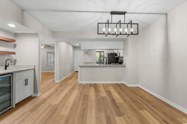 kitchen with light stone counters, ceiling fan with notable chandelier, beverage cooler, sink, and white cabinets