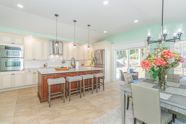 kitchen with wall chimney exhaust hood, decorative light fixtures, lofted ceiling, a kitchen island with sink, and appliances with stainless steel finishes