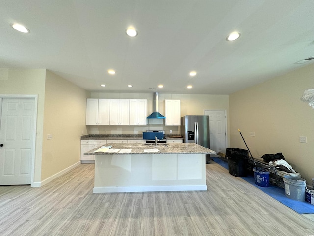 kitchen featuring stainless steel fridge, wall chimney range hood, sink, white cabinets, and an island with sink