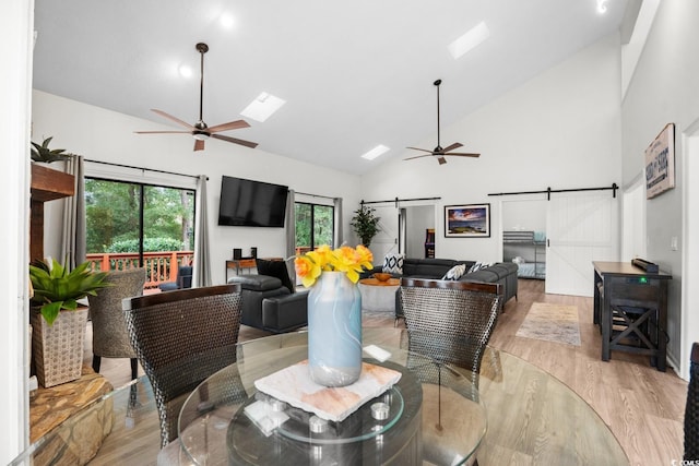 dining area with high vaulted ceiling, a skylight, ceiling fan, a barn door, and light hardwood / wood-style floors