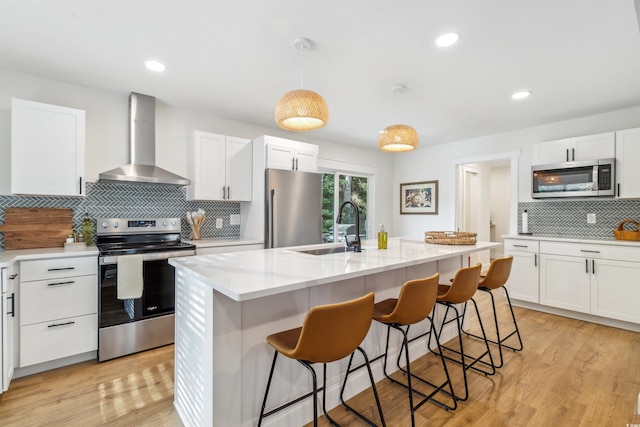 kitchen featuring sink, wall chimney exhaust hood, light wood-type flooring, an island with sink, and appliances with stainless steel finishes