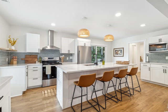 kitchen featuring wall chimney range hood, stainless steel appliances, an island with sink, and light hardwood / wood-style flooring