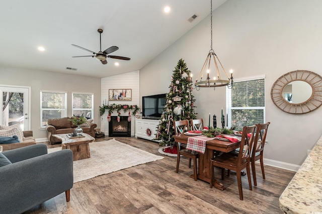 dining area featuring a large fireplace, high vaulted ceiling, wood-type flooring, and ceiling fan with notable chandelier