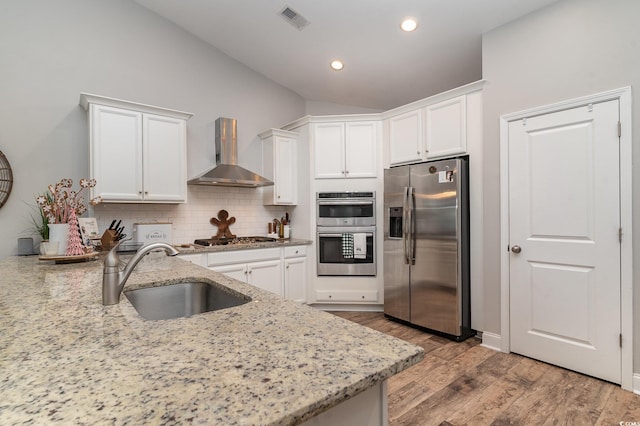 kitchen with light stone countertops, appliances with stainless steel finishes, wall chimney exhaust hood, sink, and light hardwood / wood-style flooring