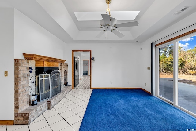 unfurnished living room featuring a skylight, a brick fireplace, a tray ceiling, ceiling fan, and light tile patterned floors