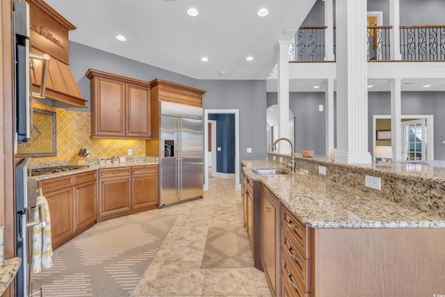 kitchen with ornate columns, light stone counters, stainless steel appliances, sink, and a large island with sink