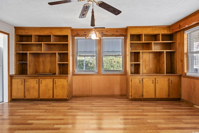 unfurnished living room featuring ceiling fan, built in features, a textured ceiling, wooden walls, and light wood-type flooring