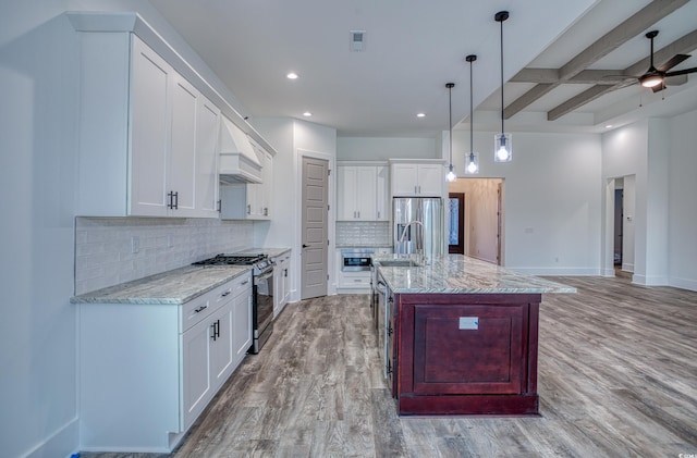 kitchen featuring beam ceiling, hanging light fixtures, an island with sink, stainless steel appliances, and white cabinets