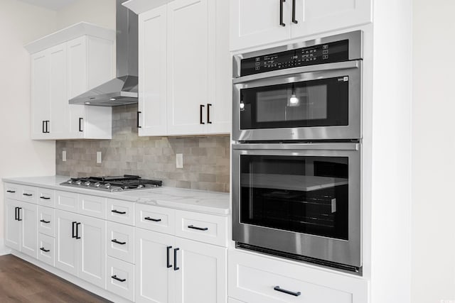 kitchen with white cabinetry, appliances with stainless steel finishes, wall chimney range hood, light stone countertops, and tasteful backsplash