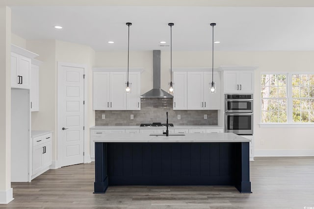 kitchen with white cabinets, an island with sink, wall chimney exhaust hood, appliances with stainless steel finishes, and hanging light fixtures